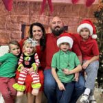 Brett McIntyre smiling with his wife Alicia and their four children in a Christmas setting, featured honoree family for Helping One Guy event.