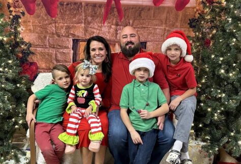 Brett McIntyre smiling with his wife Alicia and their four children in a Christmas setting, featured honoree family for Helping One Guy event.